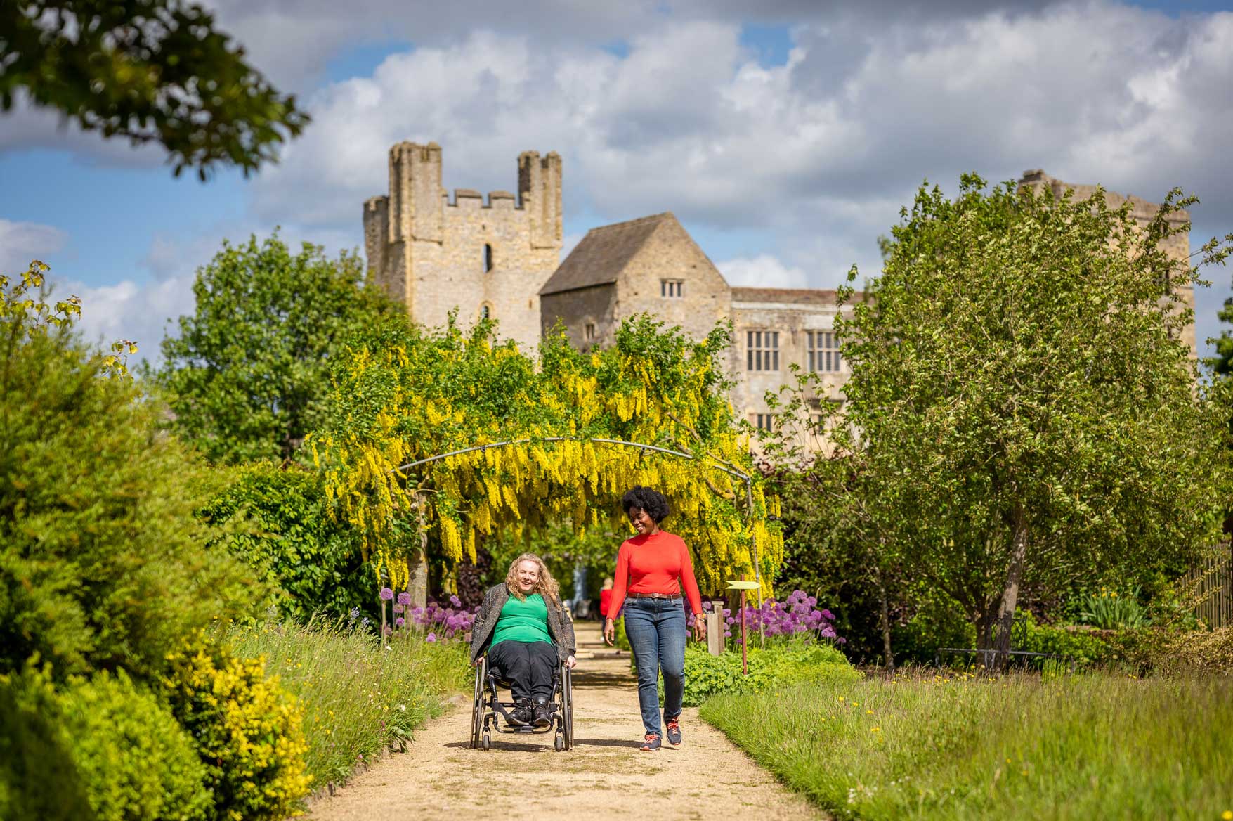 Two people, one using a wheelchair and the other walking, heading along a pathway under a flower-laden arch, with a historic castle in the background on a sunny day. Credit Visit Britain/Peter Kindersley.