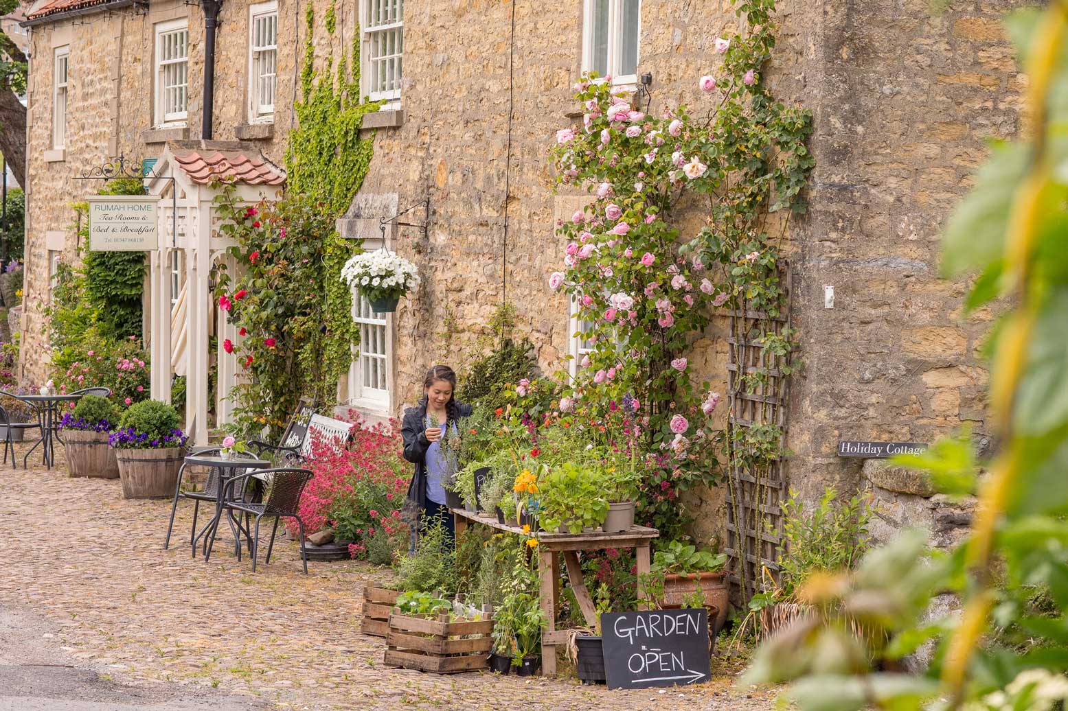 A person stands beside a rustic display of vibrant flowers and plants set up on wooden crates in a quaint stone-walled village street. A sign reading "GARDEN OPEN" is prominently displayed next to the flower arrangements. In the background, the scene is complemented by historical buildings with visible signage, lush climbing roses, and outdoor furniture, creating a peaceful and charming atmosphere. Credit Dependable Productions.