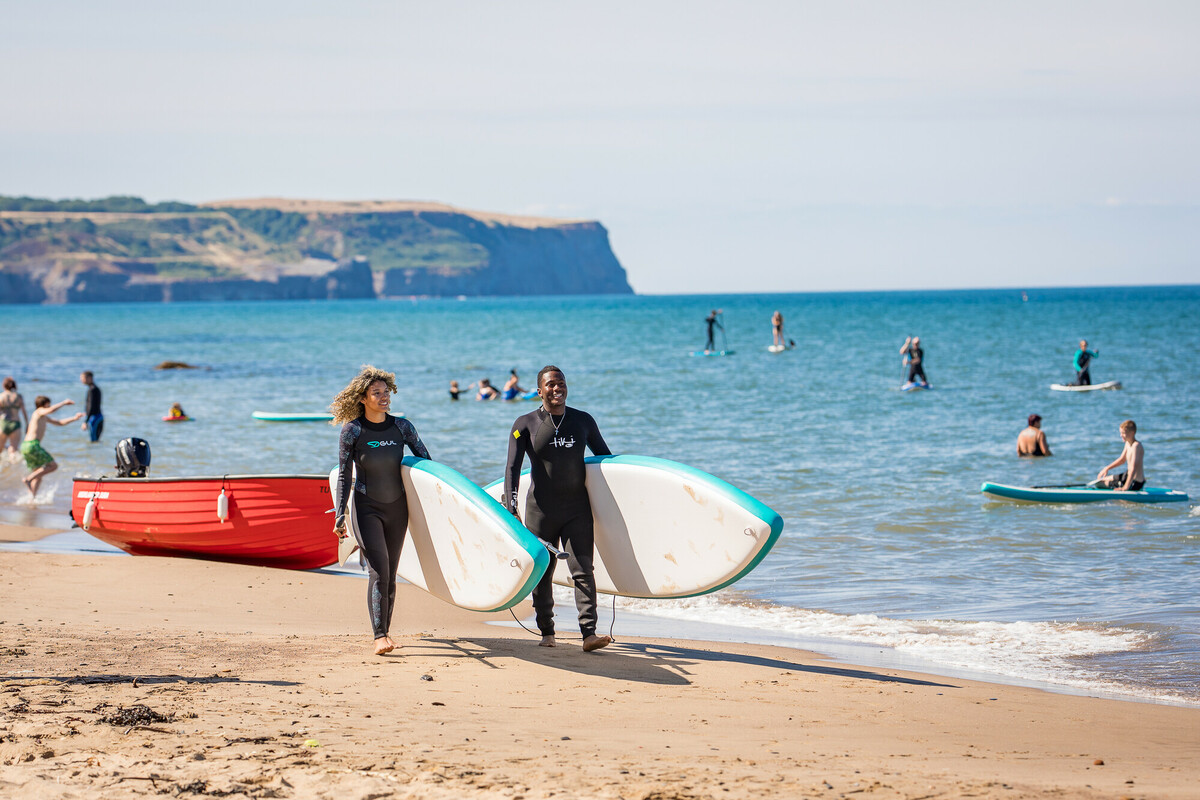 Two people carrying surfboards walk along a sandy beach with other beachgoers and paddle boarders in the water, with cliffs visible in the background. Credit Dependable Productions.