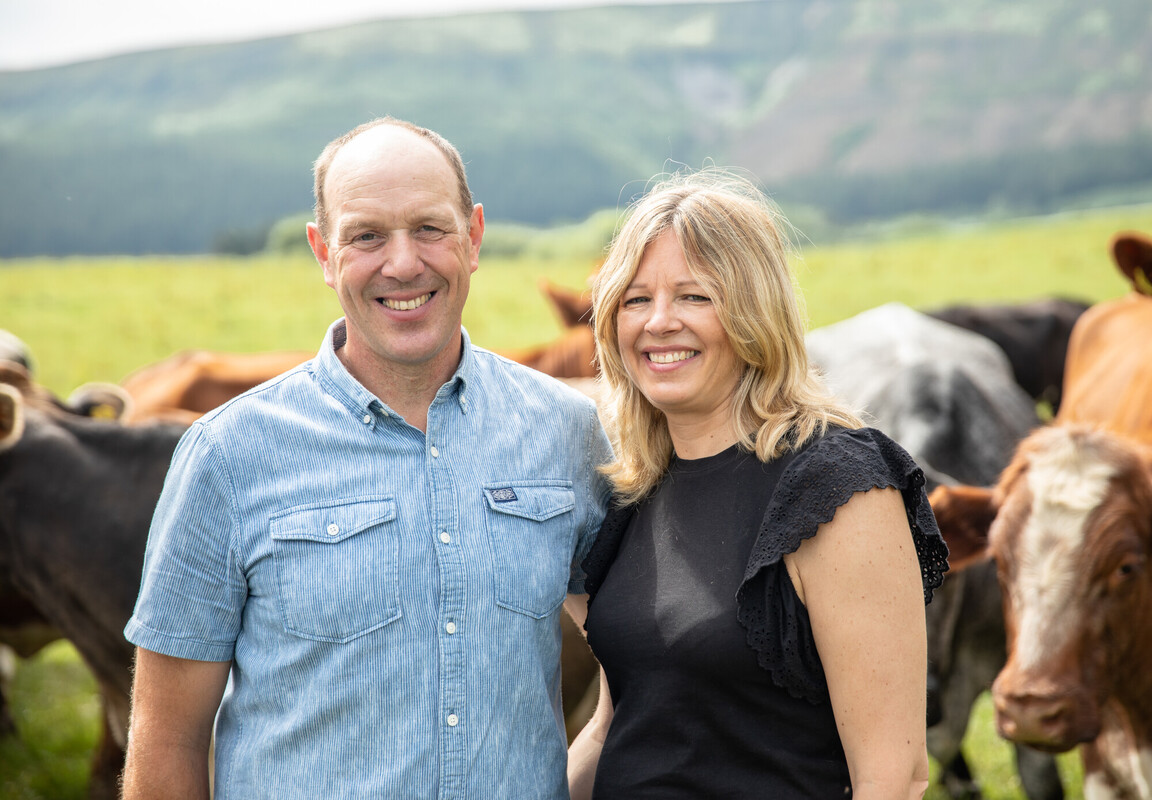 Two people with arms around each other posing for a photo. A herd of shorthorn cattle are in the background. Credit Charlie Fox.