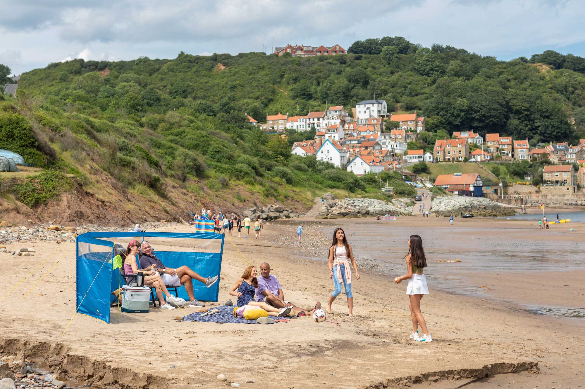 People enjoying a sunny day on a sandy beach with a backdrop of picturesque houses on a hill. Some individuals are sitting within a blue windbreak, while others are playing catch on the beach. Credit Dependable Productions.